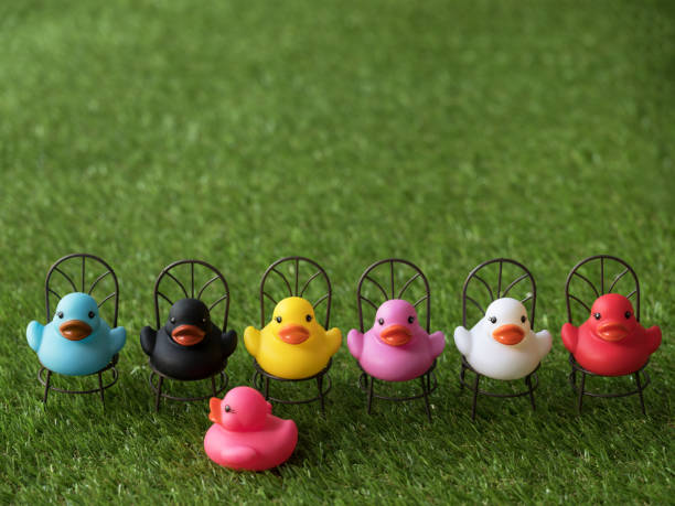 istock Line of of many different colored rubber ducks sitting on chairs facing towards the camera with one pink duck looking for a chair, scene set on artificial grass. 921680954