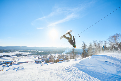 Full length action shot of young man performing snowboarding stunt spinning in air after backside flip in sunlight at ski resort, copy space