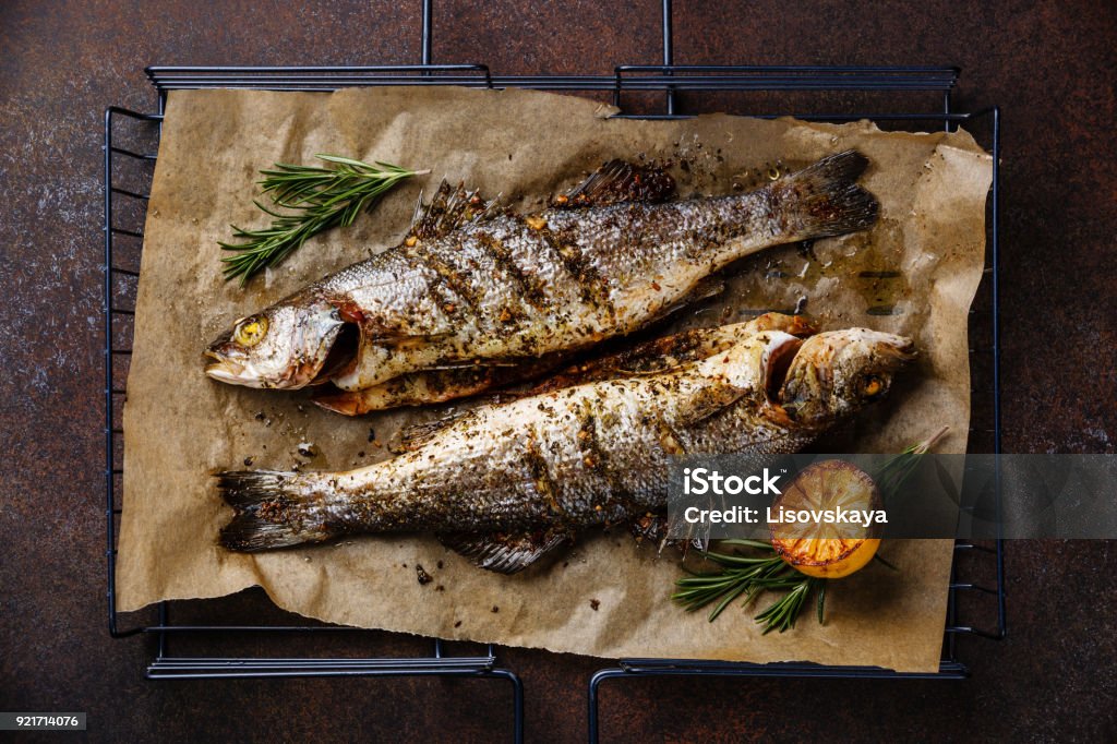 Grilled Fish Sea bass on grill with lemon and rosemary Grilled Fish Sea bass on metal grill grid with lemon and rosemary on dark background Fish Stock Photo