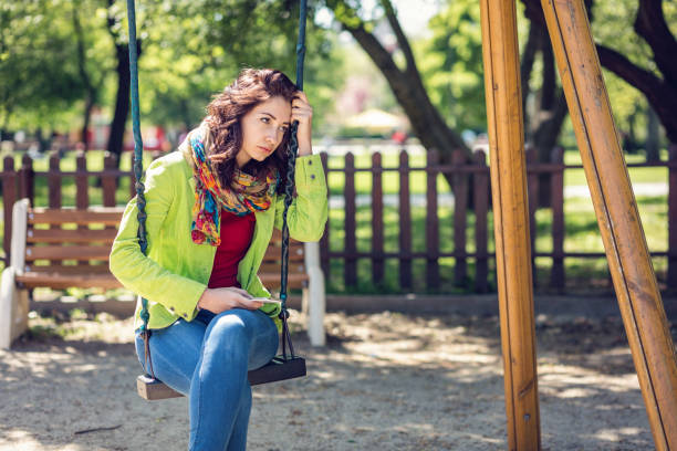 Attractive brunette female sitting on the swings at the playground and looking away unhappily