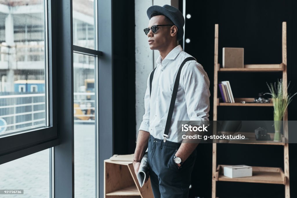 Perfect style. Handsome young man looking away and keeping hand in pocket while standing near the window Menswear Stock Photo