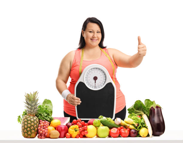 overweight woman with a weight scale making a thumb up gesture behind a table with fruit and vegetables - human hand hand sign measuring scale imagens e fotografias de stock