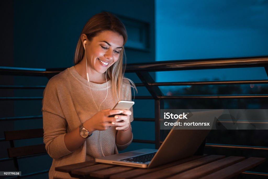 Young beautiful woman using phone and listening music at evening in her home. High ISO image. Addiction Stock Photo