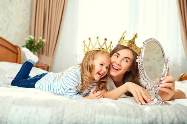Mother and daughter with crowns on the head are playing on the bed together. Mothers Day.
