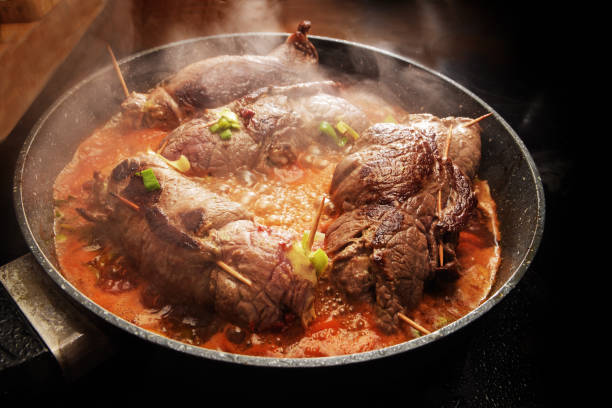 cooking beef roulades, german stuffed meat rolls with vegetables and sauce steaming in a pan on the black stove, selected focus, narrow depth of field