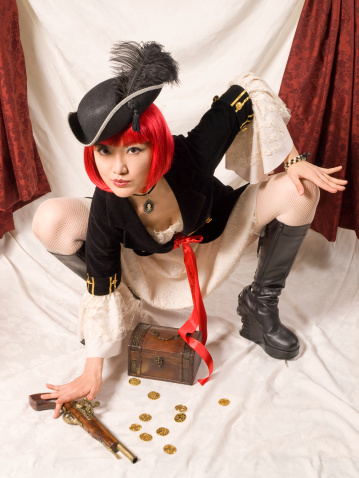 Asian woman in red bob wig and pirate costume crouching over a small chest a gold coins.