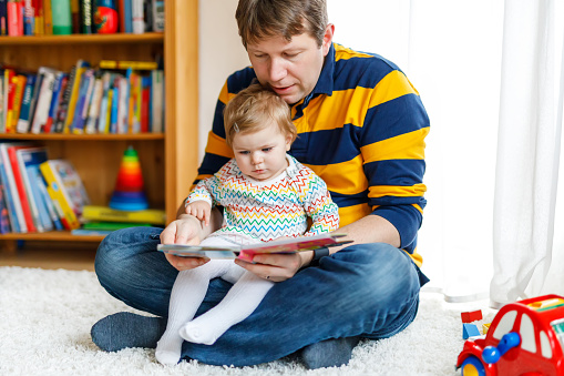 Young father reading book with his cute adorable baby daughter girl. Smiling beautiful child and man sitting together in living room at home. Toddler hearing to dad