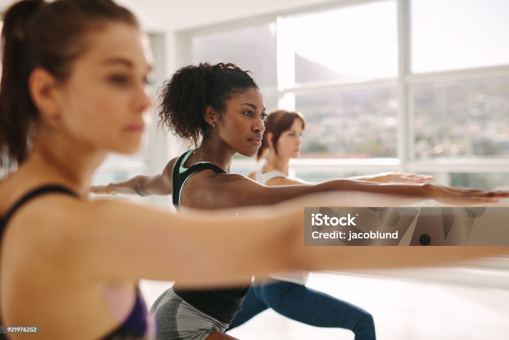 Woman doing the warrior pose during yoga class. Young woman doing the warrior pose during yoga class. Yoga instructor performing Virabhadrasana position in gym with students. Yoga Class Stock Photo