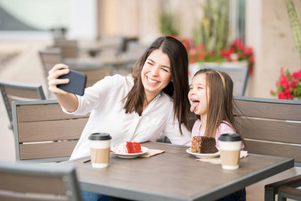 Cheerful mother and daughter at cafe taking selfie Cheerful young woman with her little girl sitting at a cafe and taking selfie with smart phone kids restaurant pictures stock pictures, royalty-free photos & images