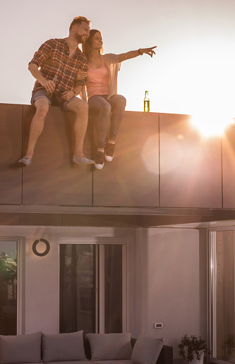 Happy couple in love having fun while relaxing on a penthouse rooftop at sunset. Woman is aiming at distance.