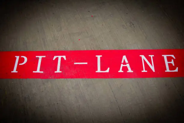 Pit-Lane is witten on a brushed concrete floor. The words are in a white, serif font on a red background and the image has a heavy vignette.