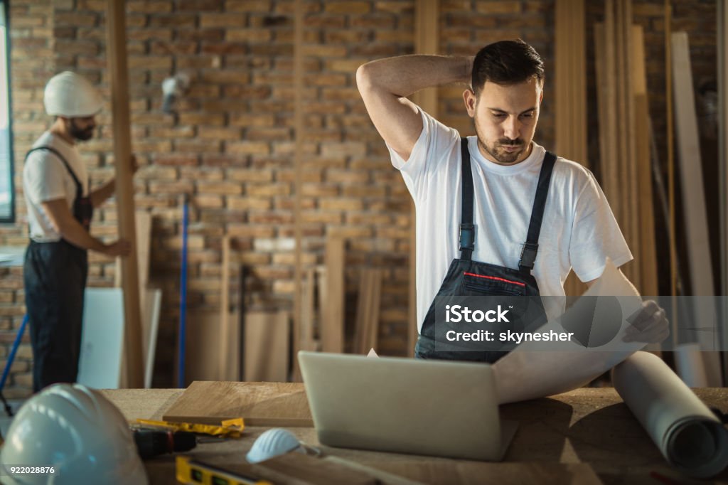 Worried manual worker having problems with blueprints at construction site. Young construction worker seeing potential problems while analyzing blueprints inside of a renovating apartment. Construction Site Stock Photo