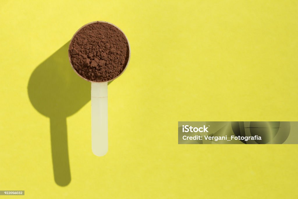 Whey Protein. Minimalism and hard light. Measuring scoop full with chocolate flavour powder on yellow background. Whey protein food supplement for training and exercise. Minimalism and hard light. Measuring scoop full with chocolate flavour powder on yellow background. Chocolate Stock Photo