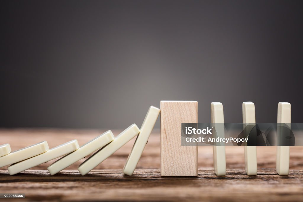 Wooden Block Amidst Falling And Upright Domino Pieces On Table Closeup of wooden block amidst falling and upright domino pieces on table against gray background Domino Stock Photo