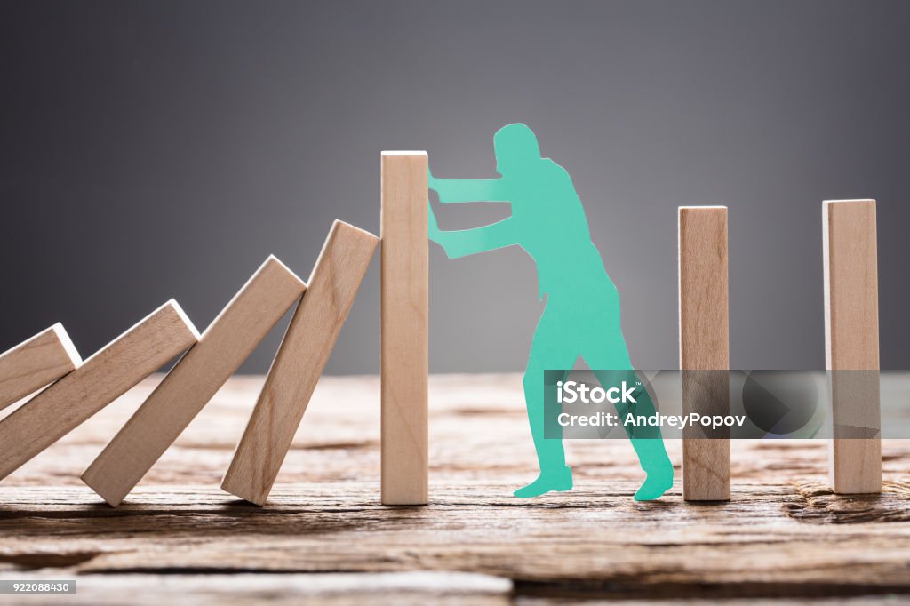 Green Paper Man Stopping Wooden Domino Blocks Closeup of green paper man stopping wooden domino blocks on table against gray background Responsibility Stock Photo