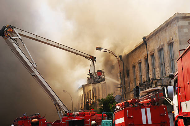 Firefighter on a firetruck, sitting on a firehose spraying water at a large blaze.