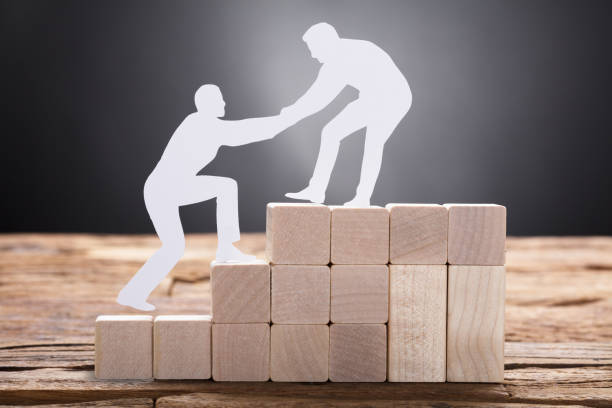 Businessman Pulling Colleague While Standing On Wooden Blocks Closeup of paper businessman pulling colleague while standing on wooden blocks against black background encouragement stock pictures, royalty-free photos & images