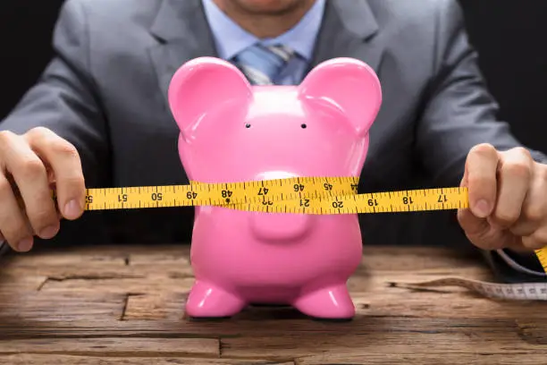 Photo of Businessman Squeezing Piggybank With Tape Measure On Table