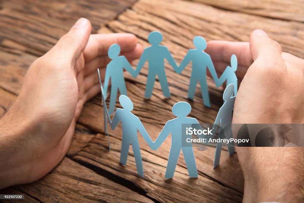 Businessman's Hands Covering Paper Team On Table Cropped image of businessman's hands covering paper team on wooden table Safety Stock Photo