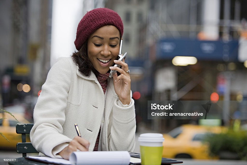 Woman at an outdoor cafe, chatting on her phone and writing An attractive business woman talking on her cell phone while seated outdoors in the city. New York City Stock Photo