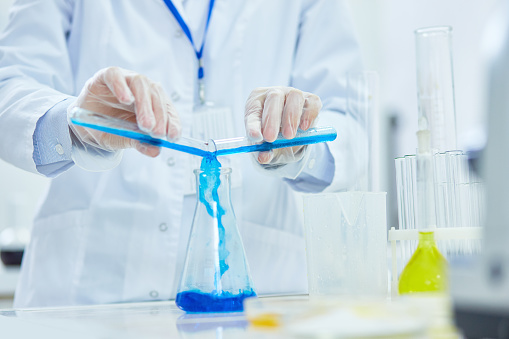 Close-up of unrecognizable chemist in lab coat pouring blue liquids in big flask while preparing for experiment in laboratory