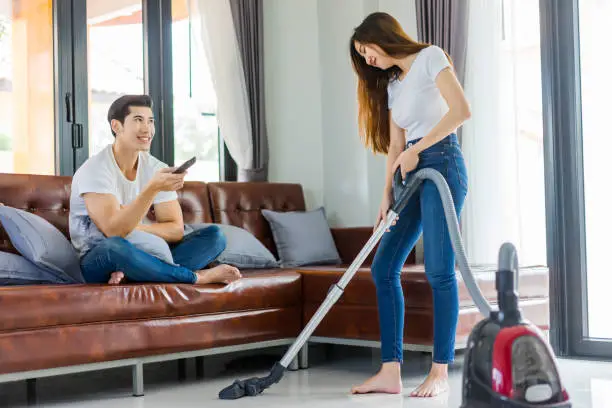 Asian couple girl doing floor cleaning with vaccuum cleaner while man on a brown sofa at home using a control remote watching television.