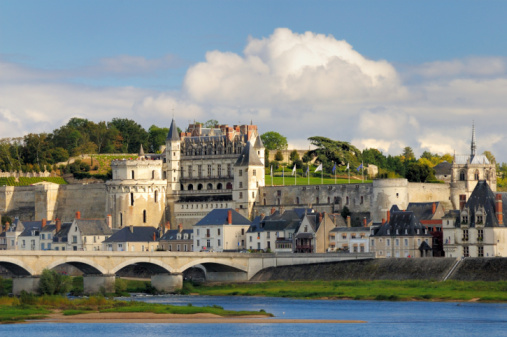 Horizontal landscape photo of the cobblestone paved walkway leading to the former drawbridge entrance in the surrounding wall of the castle at Saumur. Trees, Castle towers and rooftops can be seen in the distance under a blue sky. Saumur, Maine-et-Loire, France. 1st April, 2019