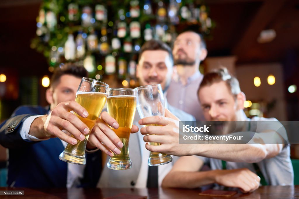 Clinking Beer Glasses Together Group of cheerful friends clinking beer glasses together while sitting at bar counter and  celebrating end of work week, focus on foreground Adult Stock Photo