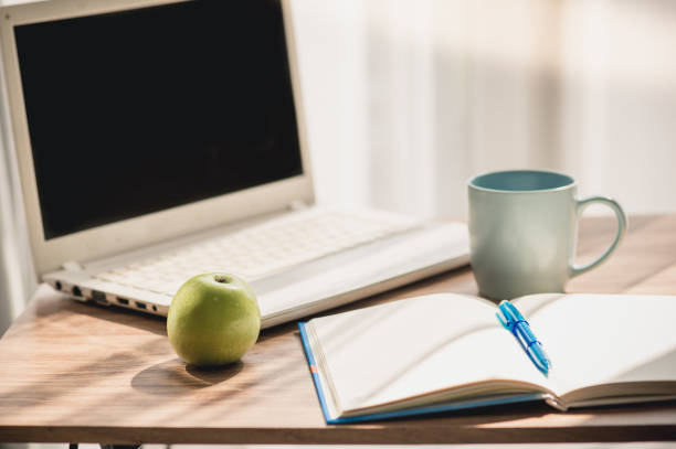 istock Green apple with laptop notebook pen and a blue coffee mug on table near window. 922271158