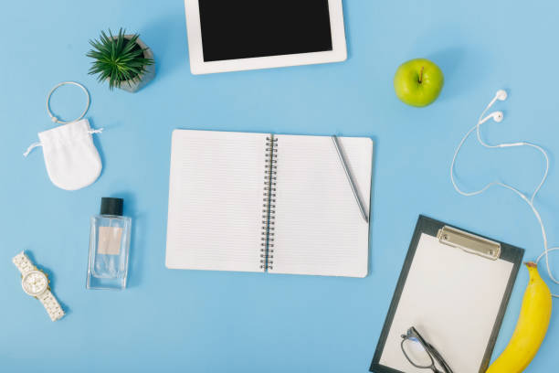 istock Workspace with  tablet, girls hands, notebook, perfumes, spectacles, fruit, headphones and felt-tip pen on blue background. Female hands write in notebook, Flat lay, top view 922289648