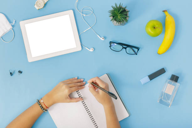 istock Workspace with  tablet, girls hands, notebook, perfumes, spectacles, fruit, headphones and felt-tip pen on blue background. Female hands write in notebook, Flat lay, top view 922289650