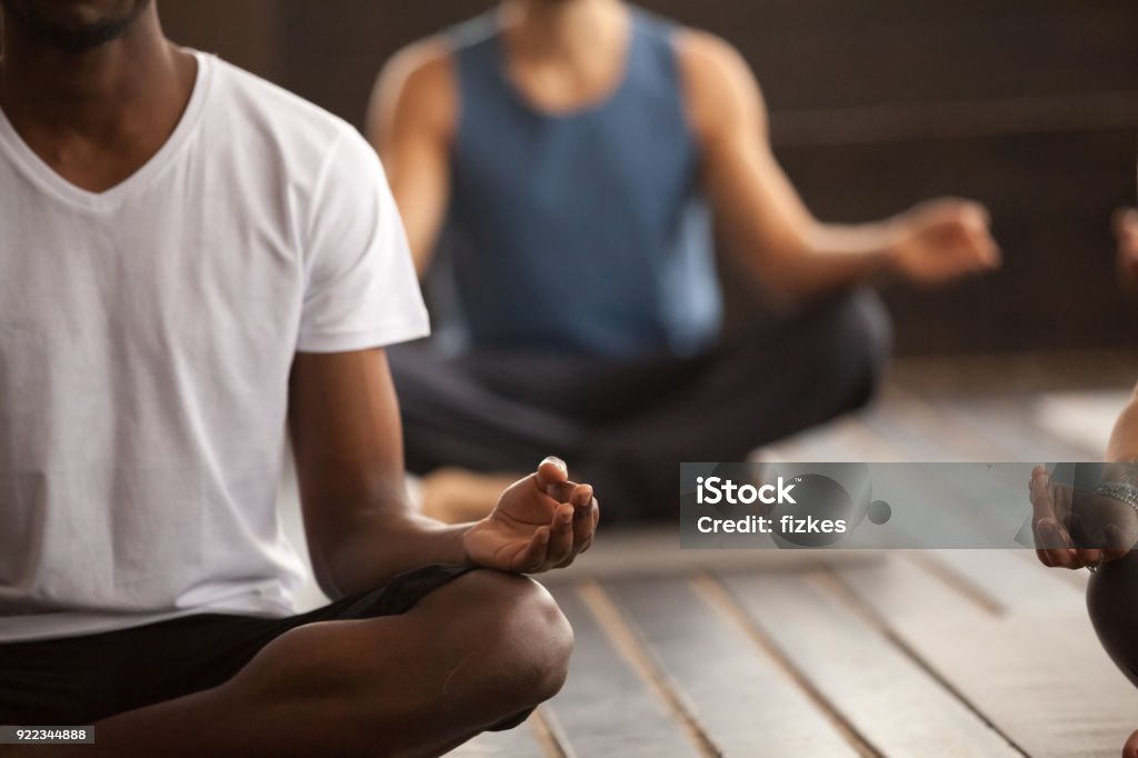 Group of sporty people in Easy Seat pose, close up Young black man and a group of young sporty people practicing yoga lesson, sitting in Sukhasana exercise, Easy Seat pose, working out, indoor close up focus on mudra gesture, studio room Yoga Stock Photo
