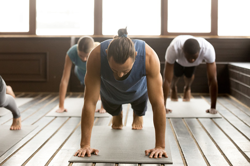 Group of young sporty people practicing yoga lesson standing in Plank pose, doing Push ups or press ups exercise, working out, indoor full length, studio