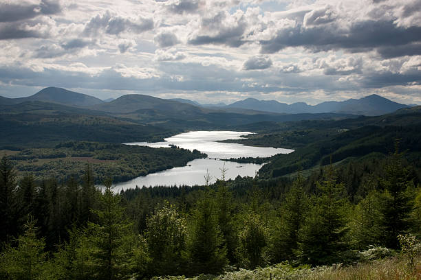 A serene view of the Scottish Highlands, with Loch Tulla surrounded by rolling green hills and dense pine forests under dramatic storm clouds.