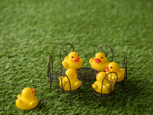 istock Group of five yellow rubber ducks sitting on chairs facing each other around a table, with one empty chair and a duck rushing towards the chair as if late for a meeting, scene set on artificial grass. 922363568