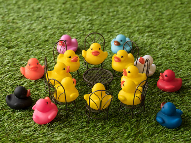 istock Group of eight yellow rubber ducks on chairs around a table facing each other, with eight different colored ducks left out of the meeting, scene set on artificial grass. 922363600