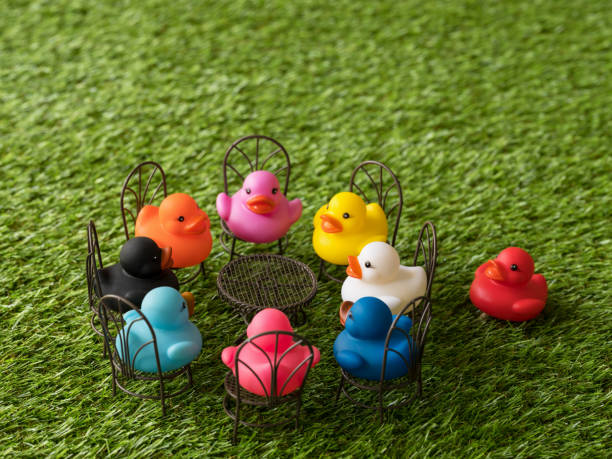 istock Group of eight different colored ducks on chairs around a table facing each other, with one red duck left out, scene set on artificial grass. 922363614