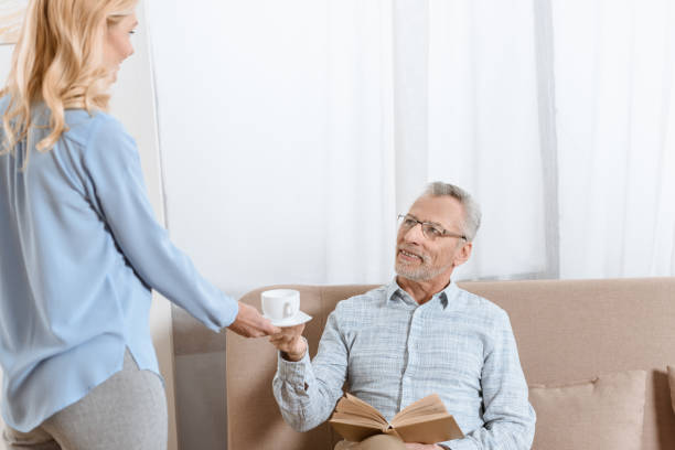 istock Mature man reading book while woman serves him tea in light room 922380456