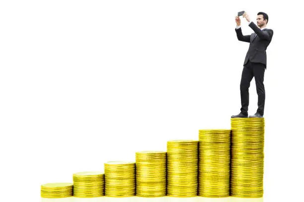 Photo of Businessman standing on top stack of coins with smart phone taking a photo on white background with copy space for business and financial concept idea.