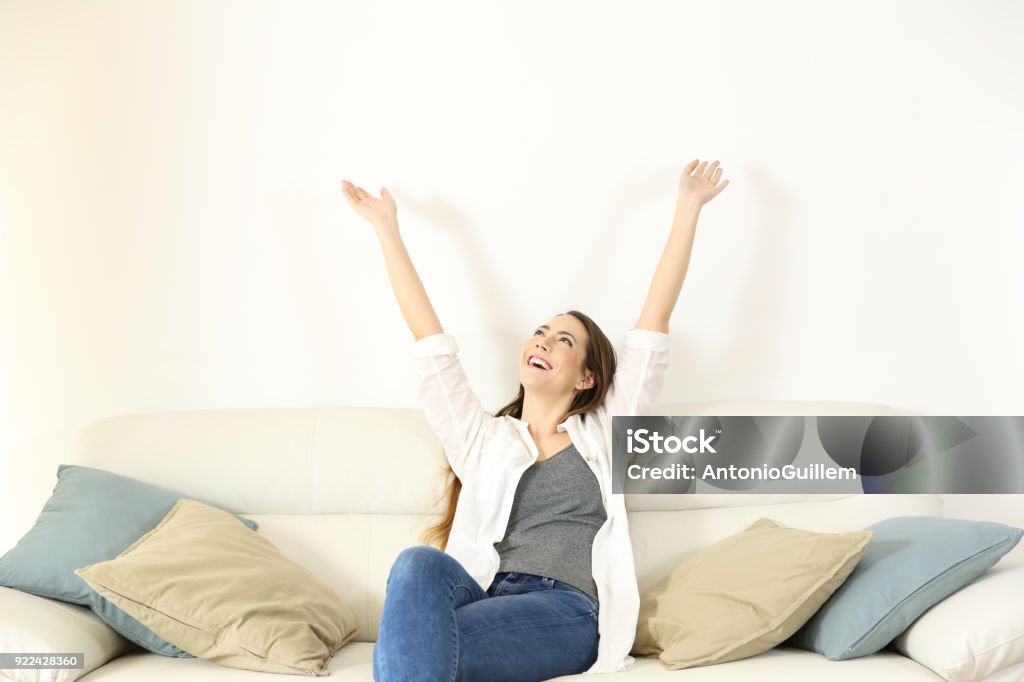 Happy woman raising arms and looking above on a couch Wide angle view portrait of a happy woman raising arms and looking above on a couch with copy space Air Conditioner Stock Photo