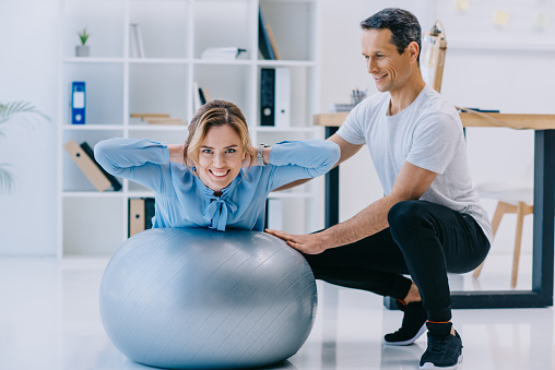 businesswoman doing hyperextension exercise on fit ball with trainer at office