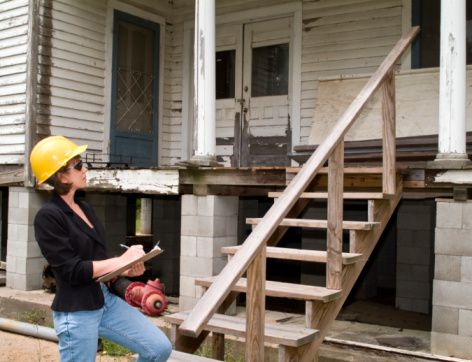 A woman in a hard hat, holding a clipboard, standing in front of an old house. 