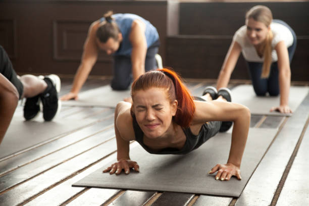 Woman doing difficult plank exercise or pushups at group training Young fit sporty woman with painful face expression doing hard difficult plank fitness exercise or push press ups feeling pain in muscles at diverse group training class in gym, endurance concept passion stock pictures, royalty-free photos & images