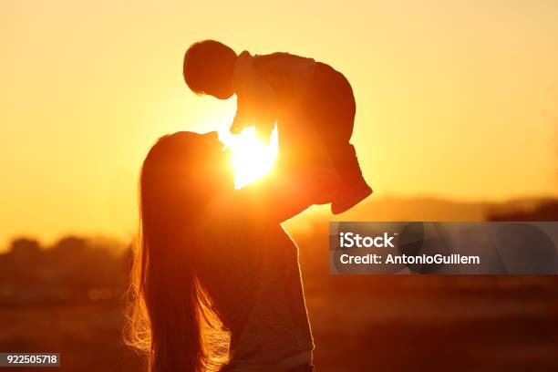 Mother Raising Her Kid Son At Sunset With The Sun In The Middle Stock Photo - Download Image Now