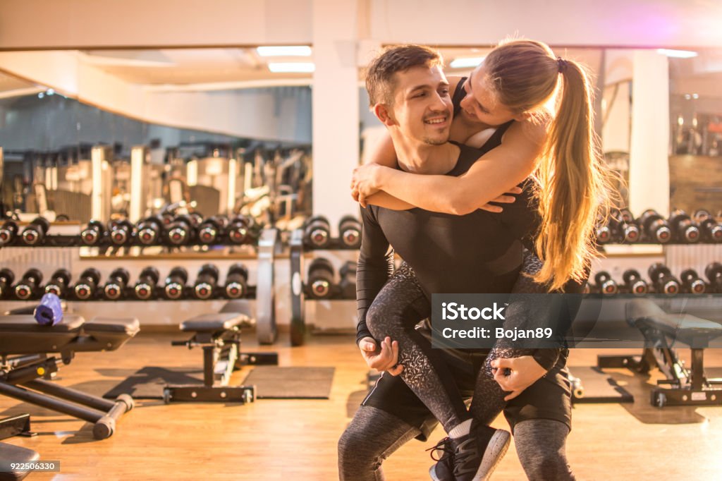 Young handsome man carrying on his girlfriend piggyback in the gym. Gym Stock Photo