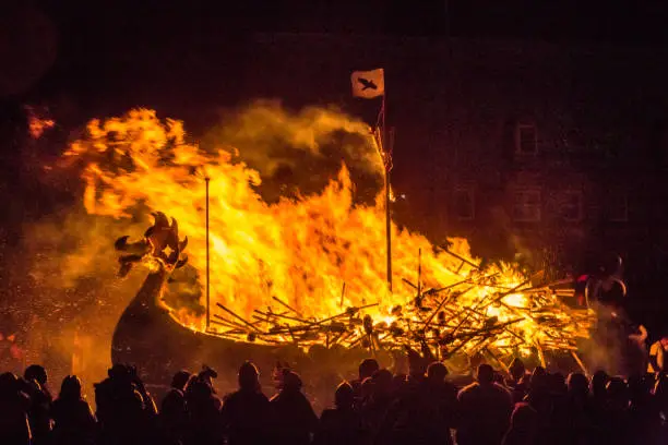 Viking longship, Blaze Away, burning at Up Helly Aa, the traditional Viking fire festival held in January. The raven flag is visible at the top of the mast, and you can see the flaming torches that have been thrown into the ship. Shadowy Viking figures are in the foreground.