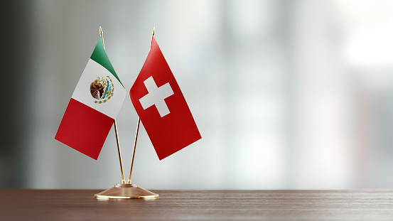 Mexican and Swiss flag pair on desk over defocused background. Horizontal composition with copy space and selective focus.