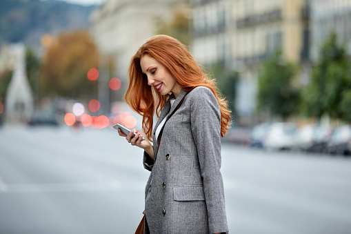 Smiling redhead woman using smart phone. Happy young female commuter walking in city. She is wearing gray blazer.