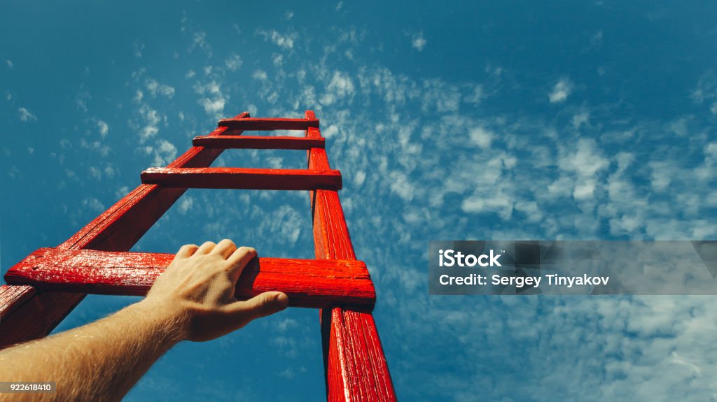 Development Attainment Motivation Career Growth Concept. Mans Hand Reaching For Red Ladder Leading To A Blue Sky A male hand holds onto the crossbar of a red wooden staircase leading to the blue sky Growth Stock Photo