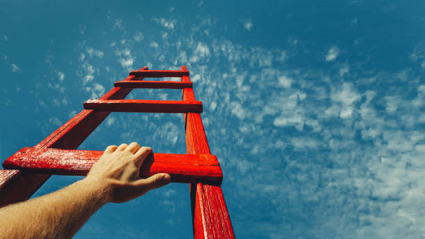 Development Attainment Motivation Career Growth Concept. Mans Hand Reaching For Red Ladder Leading To A Blue Sky A male hand holds onto the crossbar of a red wooden staircase leading to the blue sky growth stock pictures, royalty-free photos & images